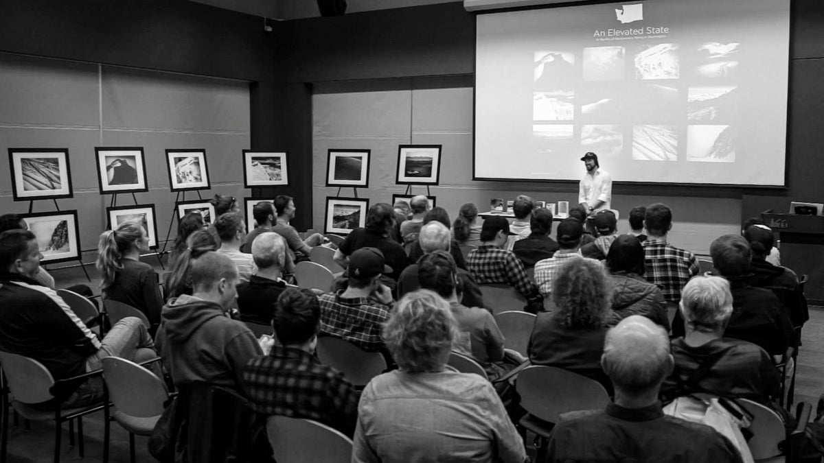 A black and white photograph of an audience attending a presentation at the Seattle REI flagship store lecture hall, with a large projection screen showing speaker Scott Rinckenberger and framed artwork displayed on easels along the left wall.